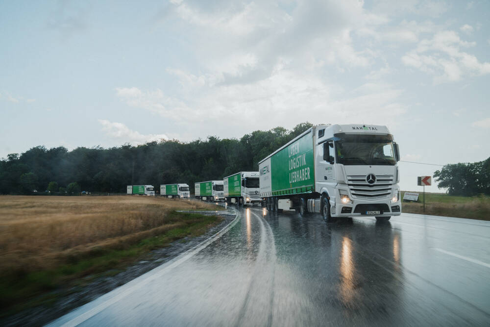 Mehrere LKWs der Firma Nahetal Logistik fahren hintereinander auf einer regennassen Landstraße.